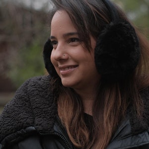 May include: A woman wearing a black puffer jacket with a fur-lined hood and black earmuffs. She is looking off to the side and smiling.