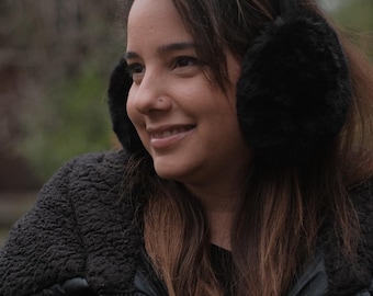 May include: A woman wearing a black puffer jacket with a fur-lined hood and black earmuffs. She is looking off to the side and smiling.