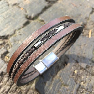 May include: A brown leather bracelet with a braided dark brown center strand and a silver-colored magnetic clasp. The bracelet has a layered design with multiple strands of leather. The bracelet is shown on a textured surface.