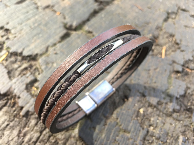 May include: A brown leather bracelet with a braided dark brown center strand and a silver-colored magnetic clasp. The bracelet has a layered design with multiple strands of leather. The bracelet is shown on a textured surface.