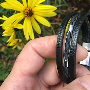 May include: Black leather bracelet with a braided center and a silver-tone clasp. The bracelet is wrapped around the wrist. Bright yellow flowers are in the background.