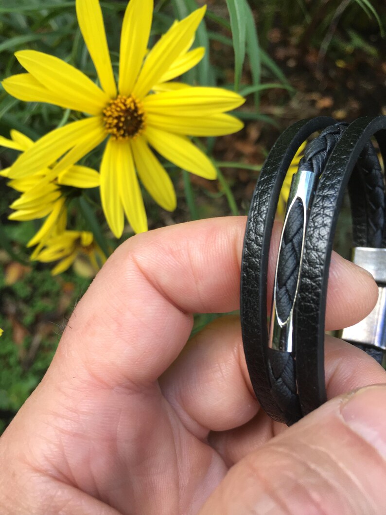 May include: Black leather bracelet with a braided center and a silver-tone clasp. The bracelet is wrapped around the wrist. Bright yellow flowers are in the background.