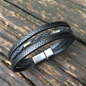 May include: Black leather bracelet with multiple strands, a braided center strand, and a silver-tone clasp. The bracelet has a textured appearance and a magnetic clasp. A stylish accessory.