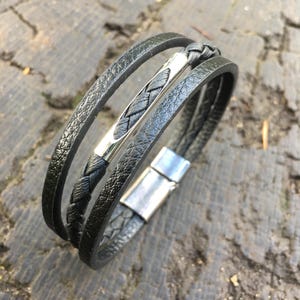 May include: Black leather bracelet with a braided center band and a silver-tone clasp. The bracelet features three bands of textured leather. The clasp is rectangular and has a magnetic closure. The bracelet is displayed on a weathered wooden surface.