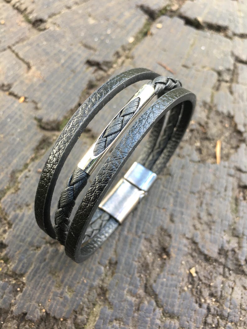 May include: Black leather bracelet with a braided center band and a silver-tone clasp. The bracelet features three bands of textured leather. The clasp is rectangular and has a magnetic closure. The bracelet is displayed on a weathered wooden surface.