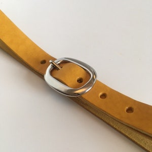 Puede incluir: Un cintur&oacute;n de cuero amarillo con una hebilla plateada. El cintur&oacute;n est&aacute; doblado por la mitad y la hebilla est&aacute; en el centro de la imagen.