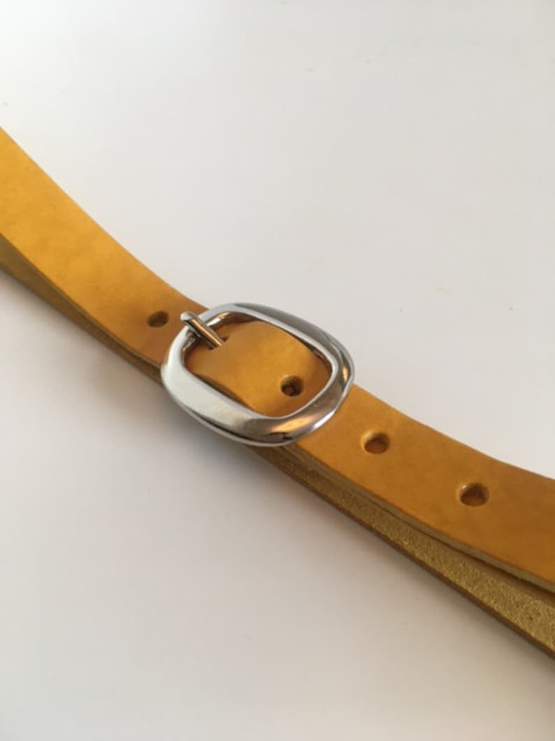 Puede incluir: Un cintur&oacute;n de cuero amarillo con una hebilla plateada. El cintur&oacute;n est&aacute; doblado por la mitad y la hebilla est&aacute; en el centro de la imagen.