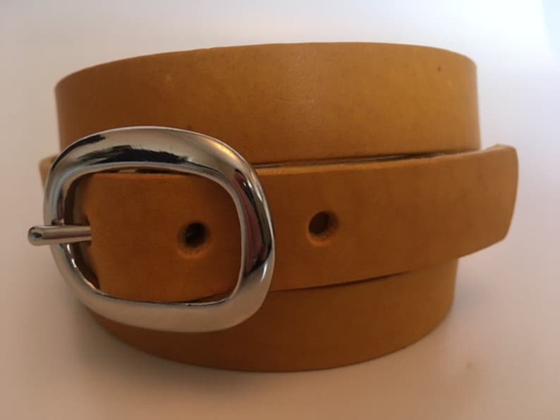 Puede incluir: Un cintur&oacute;n de cuero amarillo mostaza con una hebilla plateada. El cintur&oacute;n est&aacute; doblado por la mitad y la hebilla est&aacute; centrada en la imagen.