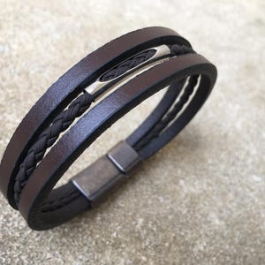 May include: Brown leather bracelet with a braided black cord and a silver-tone clasp. The bracelet features three bands of brown leather and a magnetic closure. A stylish accessory for everyday wear.
