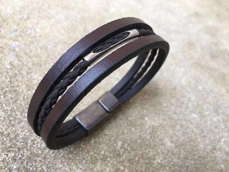 May include: Brown leather bracelet with a braided black cord and a silver-tone clasp. The bracelet features three bands of brown leather and a magnetic closure. A stylish accessory for everyday wear.