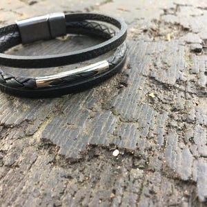 May include: A black leather bracelet with a braided band and a smooth band. The bracelet has a silver-colored clasp. The bracelet is resting on a rough, textured surface.