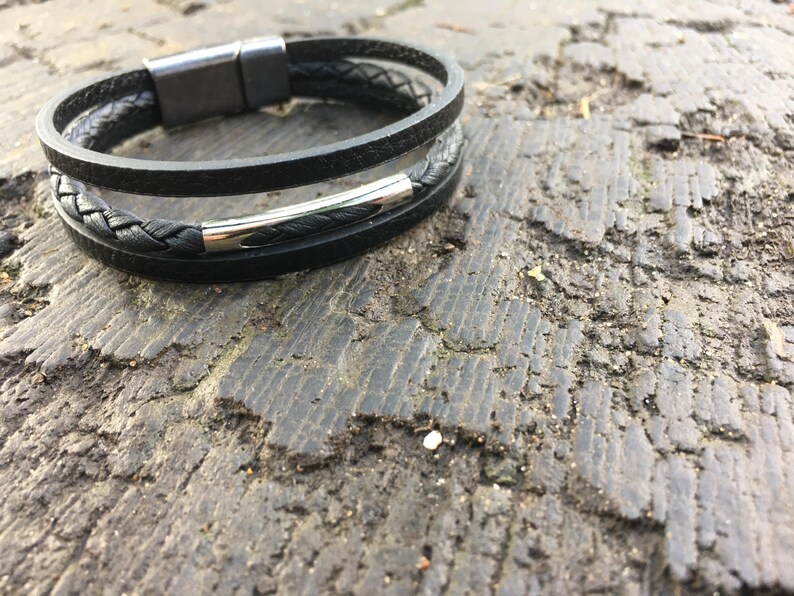 May include: A black leather bracelet with a braided band and a smooth band. The bracelet has a silver-colored clasp. The bracelet is resting on a rough, textured surface.