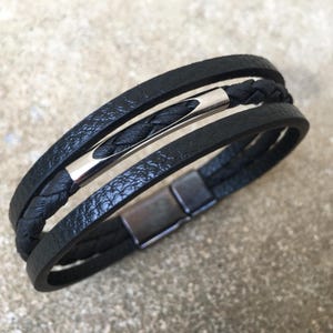 May include: Black leather bracelet with a braided center strand and a silver-tone metal bar. The bracelet has a magnetic clasp. The design features multiple layers of black leather.