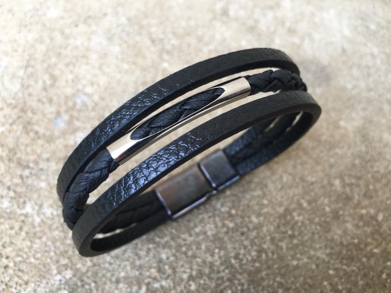 May include: Black leather bracelet with a braided center strand and a silver-tone metal bar. The bracelet has a magnetic clasp. The design features multiple layers of black leather.