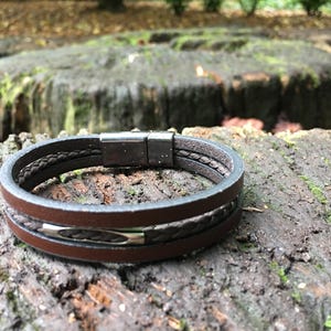May include: A brown leather bracelet with a magnetic clasp. The bracelet features a braided leather detail and a silver-tone accent. The bracelet is shown on a textured, mossy surface.