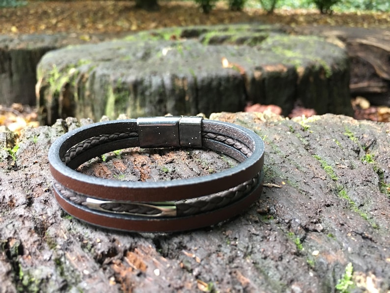 May include: A brown leather bracelet with a magnetic clasp. The bracelet features a braided leather detail and a silver-tone accent. The bracelet is shown on a textured, mossy surface.