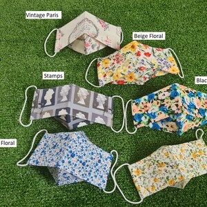 May include: Six different fabric face masks with various patterns. The masks are laid out on a green artificial grass surface. The patterns include vintage Paris, beige floral, black floral, blue floral, orange floral, and stamps.