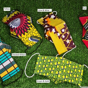 May include: Five different fabric face masks with various patterns and colors. The masks are laid out on a green artificial grass surface. The masks are labeled with their colors: Yellow, Lime & Wine, Red, Checked Ankara, and Green & Gold.