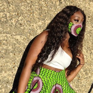 May include: A woman wearing a white crop top and a green and pink patterned skirt. She is wearing a face mask with a similar pattern.