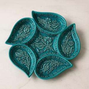 Könnte beinhalten: Eine türkisfarbene Keramikservierschale mit floralem Design. Die Schale besteht aus sechs paisleyförmigen Abschnitten, die kreisförmig angeordnet sind. Jeder Abschnitt weist ein erhabenes weißes Blumenmuster mit detaillierten Blättern und Blüten auf.