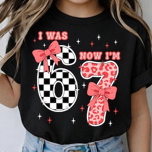 Peut inclure: T-shirt noir avec le texte "I WAS 67 NOW I'M 67". Le chiffre 6 est un motif à carreaux noir et blanc, et le chiffre 7 est un imprimé léopard rose. Les deux chiffres ont des nœuds roses.