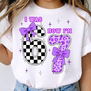 以下が含まれることがあります： 「I WAS 6 NOW I'M 7」の文字が入った白いTシャツ。数字の6は白黒の市松模様で、数字の7は紫のヒョウ柄です。どちらの数字にも紫のリボンが付いています。