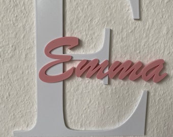 May include: White wooden letter E with the name "Emma" written in pink cursive letters on top of it.