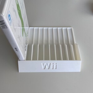 May include: White plastic stand with slots for storing video game cases. The stand has the word "Wii" printed on the front.