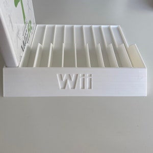 Puede incluir: Soporte de plástico blanco para cajas de juegos con ranuras para múltiples cajas de juegos. El soporte tiene la palabra "Wii" impresa en la parte delantera.