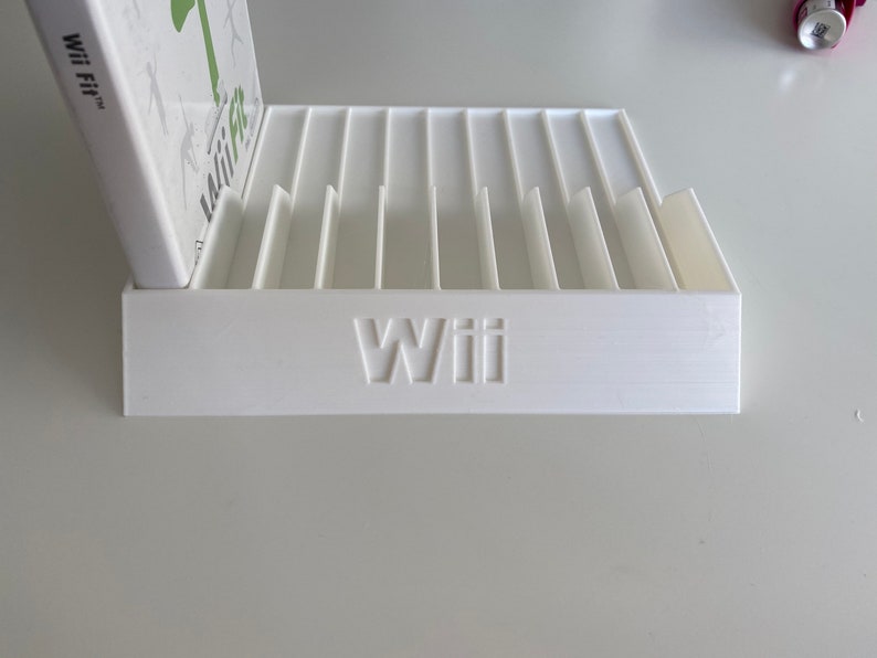 May include: White plastic game case holder with slots for multiple game cases. The holder has the word "Wii" printed on the front.
