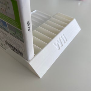 May include: White plastic video game storage stand with slots for game cases. The stand has the word "Wii" printed on the front. A white video game case with the text "Wii Fit" is resting on the stand.