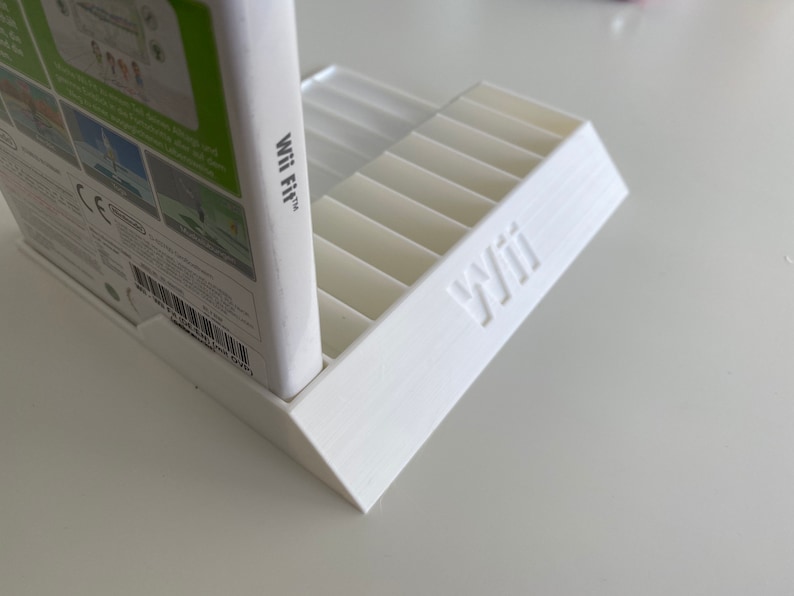 May include: White plastic video game storage stand with slots for game cases. The stand has the word "Wii" printed on the front. A white video game case with the text "Wii Fit" is resting on the stand.