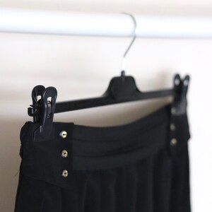May include: Black pleated skirt with a high waist and gold button accents hanging on a black hanger.