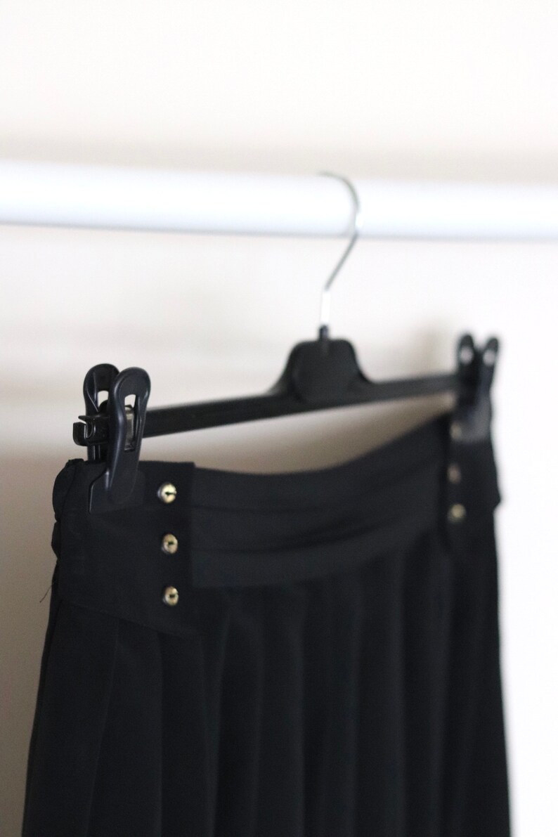 May include: Black pleated skirt with a high waist and gold button accents hanging on a black hanger.
