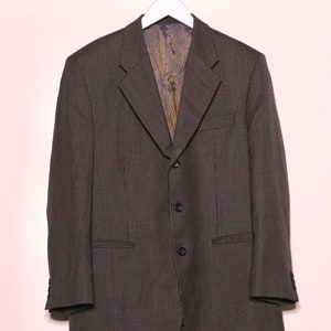 Könnte beinhalten: Ein dunkelgrauer Blazer mit einem dezenten Karomuster hängt an einem weißen Kleiderbügel. Die Dreiknopfjacke hat zwei Vordertaschen und ein fallendes Revers. Das Innenfutter ist goldfarben.
