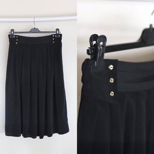 May include: A black pleated skirt with gold button accents. The skirt is knee-length and has a straight silhouette.