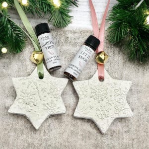 May include: Two white star-shaped Christmas decorations with snowflake patterns, suspended by ribbons and small gold bells. Two essential oil bottles are positioned nearby. One reads "CHRISTMAS TREE" and the other "LET IT SNOW".