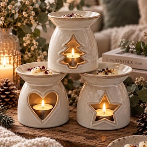 May include: Three cream-coloured ceramic wax warmers with a glossy finish and brown-toned edges. Each warmer features a cutout shape: a Christmas tree, a heart, and a star. A lit tea light candle sits inside each base, and wax melts are in the top bowls.