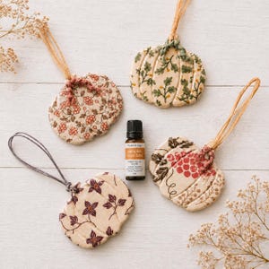 May include: Four ceramic pumpkin-shaped ornaments with various floral patterns in shades of red, green, and brown, each featuring a natural fibre hanging loop. A small amber glass bottle of Pumpkin Spice Latte essential oil sits in the centre.