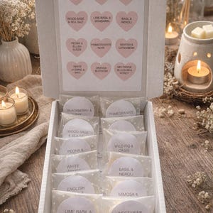 May include: A white box filled with scented wax melts, each labeled with a different fragrance name like "Blackberry & Bay" and "Lime Basil & Mandarin." The box is open, revealing the individual melts in clear packaging. The background includes candles and floral arrangements.