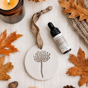May include: A round white ceramic ornament featuring an embossed dandelion design, suspended by a patterned brown ribbon, accompanied by a 5ml amber glass dropper bottle labelled Flicker & Scent calm essential oil blend with bergamot and lavender.