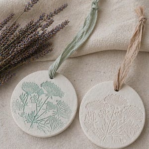 May include: Two circular ceramic ornaments with floral patterns and ribbons. One has a light green floral design, the other an embossed design. A bundle of lavender is visible in the background. Each ornament is approximately 8 cm in diameter.