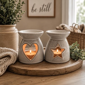 May include: Two glossy, light grey ceramic wax warmers with a smooth finish. One warmer features a heart-shaped cutout, and the other features a star-shaped cutout. Each warmer contains a small, lit tea light candle inside the central opening.
