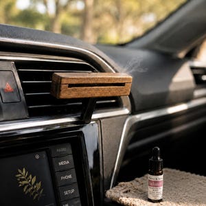 May include: A rectangular wooden car air freshener clip attached to a vehicle air vent, emitting a faint wisp of vapour. Beside it sits a small amber glass dropper bottle with a white label reading Wicker & Sage Balance Essential Oil Blend.