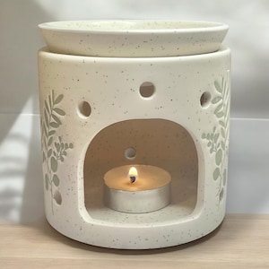 May include: White ceramic oil diffuser with a leafy design and a small candle inside. The diffuser has a top opening for adding essential oils.