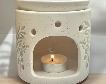 May include: White ceramic oil diffuser with a leafy design and a small candle inside. The diffuser has a top opening for adding essential oils.