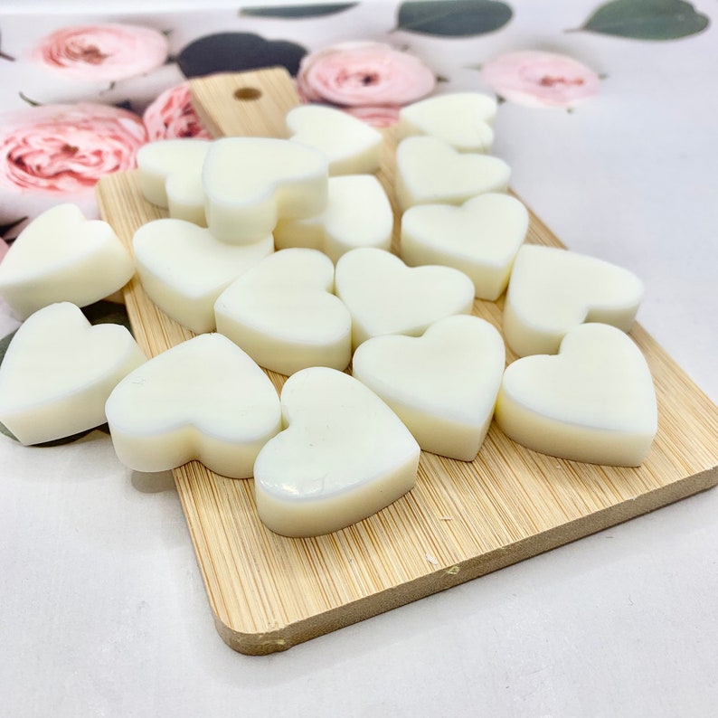 May include: A collection of heart-shaped wax melts in a creamy white color, arranged on a small, light-brown wooden cutting board. The melts are smooth and have a glossy finish. The background features a floral pattern.
