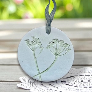 May include: A circular, pale blue ornament featuring an embossed floral pattern. It has a dark grey ribbon for hanging. The background is blurred, with a white lace doily in the foreground.