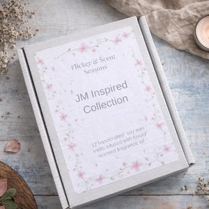 May include: A rectangular box with the text "JM Inspired Collection" and floral accents. The box is on a light blue wooden surface, with a lit candle in a silver holder and dried flowers nearby. The box contains 12 handcrafted soy wax melts.