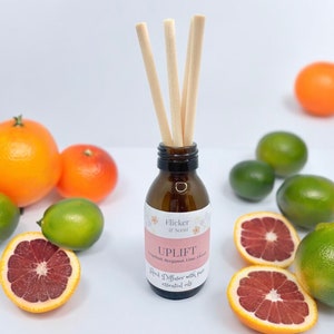May include: A brown glass bottle diffuser with wooden sticks and a pink label that reads "Flicker & Scent" and "UPLIFT Grapefruit, Bergamot, Lime, Orange Reed Diffuser with pure essential oils". The bottle is surrounded by limes and oranges.